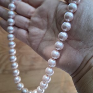May include: A single strand necklace featuring a series of uniform, round, light pink pearls. The pearls have a smooth, lustrous finish and are strung together in a continuous line, displaying a soft, reflective sheen across their spherical surfaces.