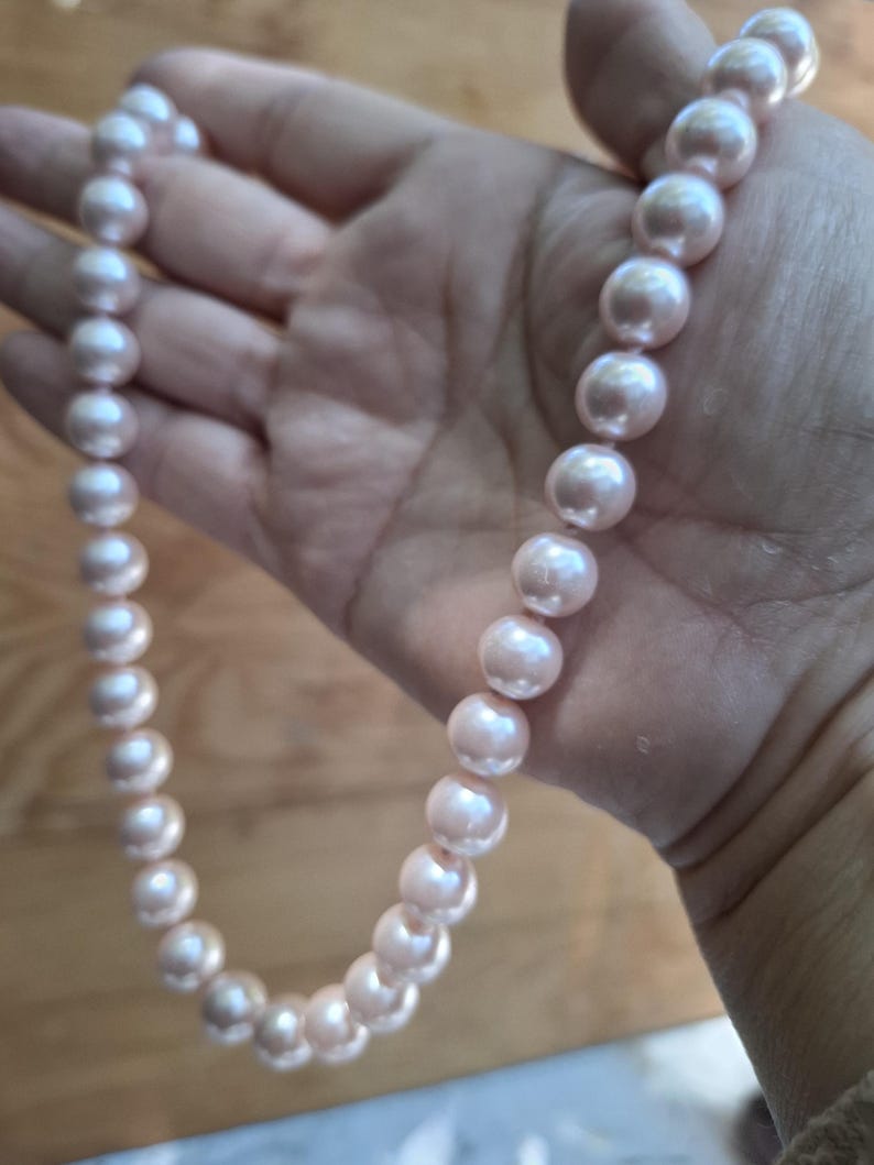 May include: A single strand necklace featuring a series of uniform, round, light pink pearls. The pearls have a smooth, lustrous finish and are strung together in a continuous line, displaying a soft, reflective sheen across their spherical surfaces.