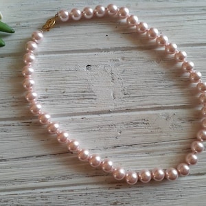 May include: A single strand necklace featuring round, light pink pearls of uniform size. The necklace is secured with a gold-toned metal clasp that connects the two ends of the pearl strand, creating a circular shape for the jewellery piece.