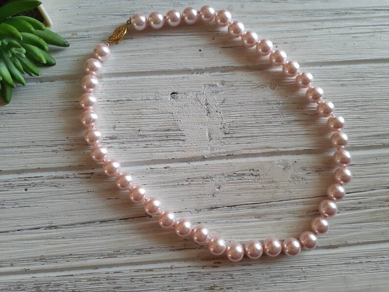 May include: A single strand necklace featuring round, light pink pearls of uniform size. The necklace is secured with a gold-toned metal clasp that connects the two ends of the pearl strand, creating a circular shape for the jewellery piece.