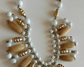 May include: A statement necklace featuring a central chain adorned with alternating white spherical beads and metallic gold-toned beads. The lower section displays a fringe of elongated, ribbed gold-toned beads interspersed with smaller white beads.