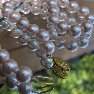 May include: A strand of lustrous, light pink spherical beads arranged in a coiled pattern. The necklace features a gold-toned metal clasp with an oval, textured design and a hook-style closure, reflecting light across the smooth, round surfaces.