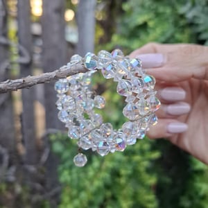 May include: A multi-strand wrap bracelet composed of clear, faceted crystal beads with an iridescent finish. The bracelet features a coiled design with multiple rows of beads and a single small crystal bead dangling from one end of the wire structure.