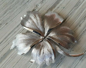 May include: A silver-toned metal brooch shaped like a five-petaled flower. The petals feature a brushed, textured finish with smooth, polished edges that catch the light. The design is symmetrical with overlapping petals radiating from the centre point.