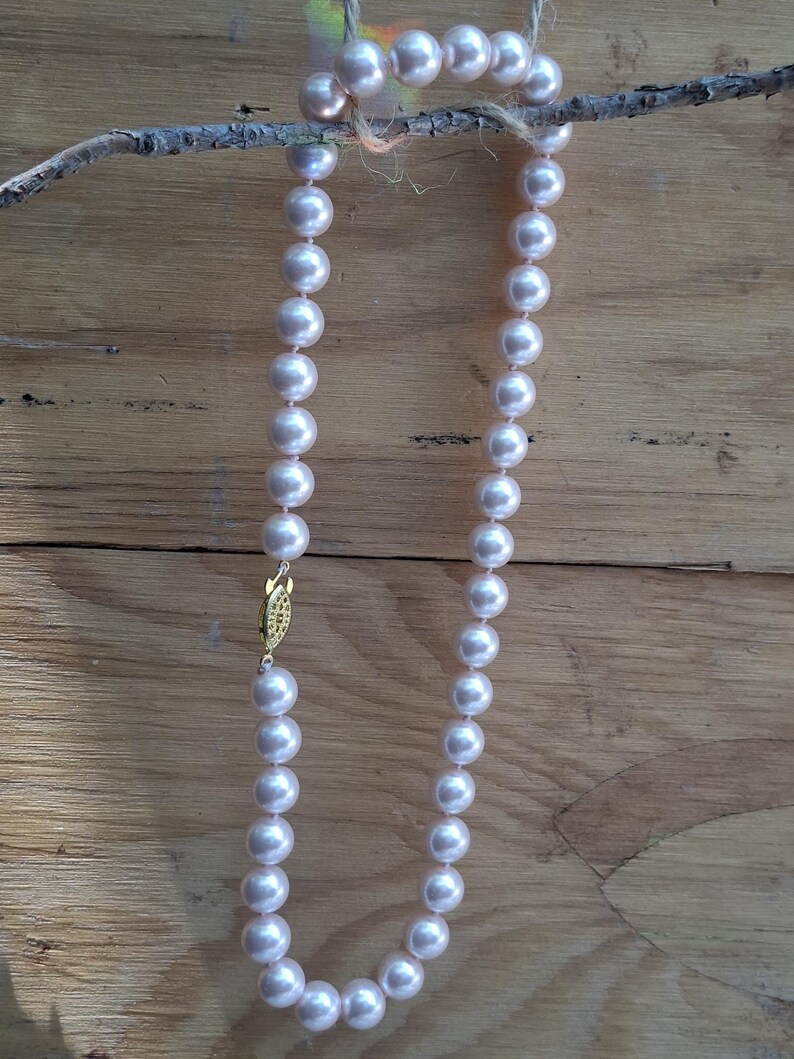 May include: A single strand necklace featuring uniform, round, pale pink pearls with a lustrous finish. The necklace is secured with a decorative, oval-shaped gold-tone metal clasp that features intricate engraved detailing on its surface.