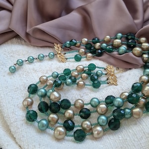 Peut inclure: Un collier multi-rangs composé de perles alternées dans des tons vert émeraude, bleu sarcelle et or champagne. Le design comprend des perles vertes translucides à facettes et des perles rondes lisses, reliées par des fermoirs en métal doré.