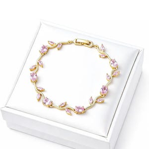 May include: A gold-toned metal bracelet featuring a vine-like design with alternating oval-shaped pink gemstones and small leaf-shaped accents. The jewelry piece includes a rectangular clasp mechanism and follows a circular, flexible link structure.