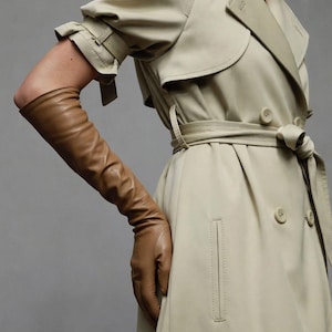 May include: A beige double-breasted trench coat with short sleeves featuring buckled cuffs, a waist belt, and side pockets, paired with long, tan leather opera gloves that extend past the elbow, creating a layered look.