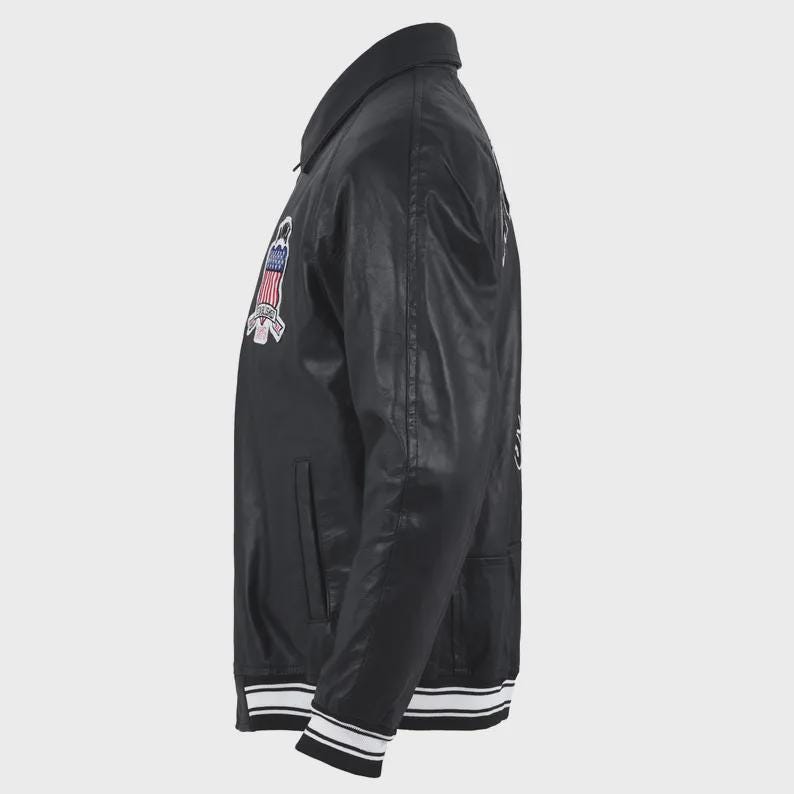 May include: A black jacket with a fold-down collar, long sleeves, and ribbed cuffs featuring white stripes. The chest displays a patch with a red, white, and blue shield design. The jacket has a front zipper closure and two side pockets.