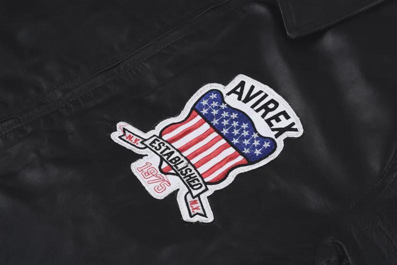May include: A black leather jacket featuring an embroidered patch on the chest. The patch displays the text AVIREX, ESTABLISHED 1975, and N.Y. along with a graphic of the American flag shaped like a shield with red, white, and blue colors.
