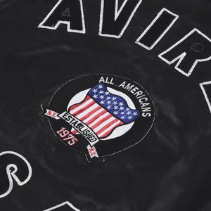 May include: A black jacket featuring the white embroidered text AVIREX and U.S. along with a circular patch. The patch displays a United States flag, the text ALL AMERICANS, ESTABLISHED 1975, and N.Y. in white and red embroidery on a black background.