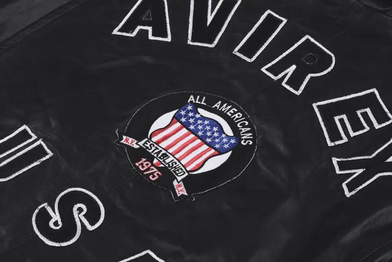 May include: A black jacket featuring the white embroidered text AVIREX and U.S. along with a circular patch. The patch displays a United States flag, the text ALL AMERICANS, ESTABLISHED 1975, and N.Y. in white and red embroidery on a black background.