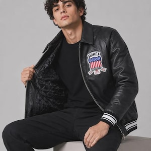 May include: A black leather jacket featuring a front zipper, a collar, and ribbed cuffs with white stripes. A patch on the chest displays the text AVIREX above a shield graphic with red and white stripes and blue stars, with the text ESTABLISHED 1975.