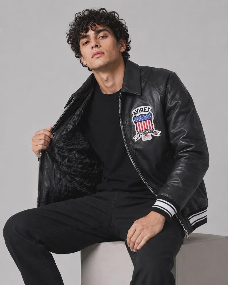 May include: A black leather jacket featuring a front zipper, a collar, and ribbed cuffs with white stripes. A patch on the chest displays the text AVIREX above a shield graphic with red and white stripes and blue stars, with the text ESTABLISHED 1975.