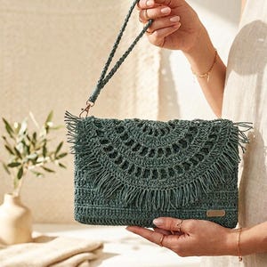 May include: A dark green crochet clutch bag featuring a semi-circular flap with fringe detailing and a matching braided wrist strap. The rectangular bag includes a small metallic rectangular tag on the bottom right corner and a gold-toned metal clasp.