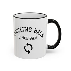 May include: A white ceramic mug with a black handle and black rim. The front features the black text "CIRCLING BACK SINCE 9AM" in a bold, sans-serif font, positioned above a black circular arrow icon representing a refresh or reload symbol.