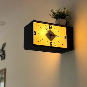 May include: A rectangular wall-mounted clock with a black frame and a glowing yellow face. The clock features black numerals at the 12, 3, 6, and 9 positions, black hour and minute hands, and a small potted plant sitting on top of the housing.