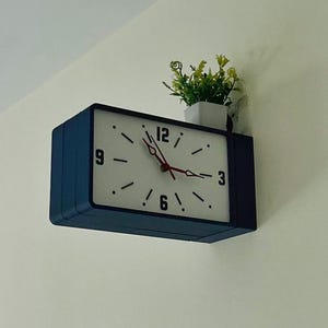 May include: A rectangular wall clock with a dark blue frame and a white face. The clock features black numerals at the 3, 6, 9, and 12 positions, with black tick marks for other hours. It has red hour and minute hands and a small potted plant on top.