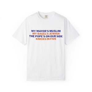 Op de afbeelding: Een wit t-shirt met korte mouwen en de tekst in blauw en oranje: MY MAYOR'S MUSLIM, MY BAGEL'S JEWISH, THE POPE'S ON OUR SIDE, KNICKS IN FIVE. Het shirt heeft een ronde hals en een standaard pasvorm met de tekst duidelijk weergegeven.