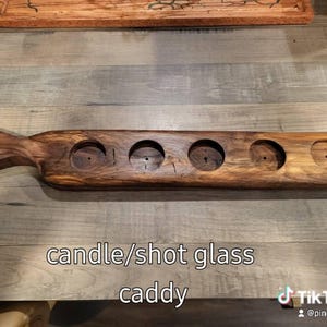 May include: A long, dark brown wooden serving board featuring a handle on one end and five circular cutouts designed to hold items. The text candle/shot glass caddy is printed on the image. The wood grain is visible throughout the entire piece.