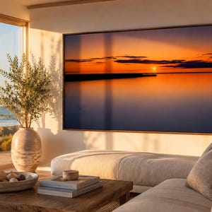 May include: A large rectangular wall art piece featuring a vibrant sunset over a calm body of water with orange and deep blue hues, framed in a thin dark wood border, displayed on a light-colored wall above a beige sofa and wooden coffee table.