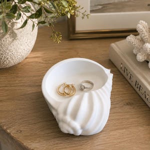 May include: A white ceramic trinket dish shaped like a seashell with a ribbed exterior. The interior holds a pair of gold hoop earrings and a silver ring with a clear stone. The dish has a smooth matte finish and a curved, open design.
