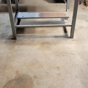 May include: A gray metal workbench featuring a flat rectangular top surface, four sturdy vertical legs, and a lower horizontal shelf with visible rust discoloration. The frame is constructed from square metal tubing with bolted corner joints.
