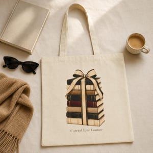 May include: A cream-colored canvas tote bag featuring a graphic illustration of a stack of books tied with a ribbon bow. The text "Carried Like Couture" is printed in black below the illustration. The bag has two long shoulder straps.