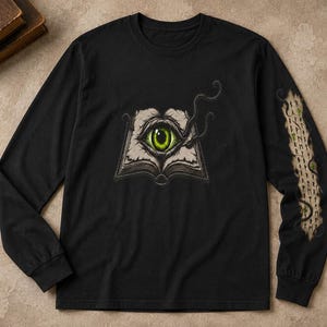 Puede incluir: Una camiseta negra de manga larga con el gráfico de un libro abierto que muestra un ojo verde grande y detallado en el centro. La manga derecha presenta una franja vertical con símbolos rúnicos y un diseño de humo oscuro y ondulante.