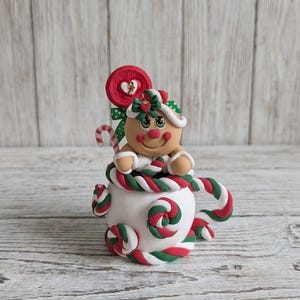 May include: A clay figurine of a gingerbread person sitting inside a white mug decorated with red and green candy cane swirls. The figure features a red lollipop, a green bow, and a small candy cane. The mug is adorned with multiple candy cane accents.
