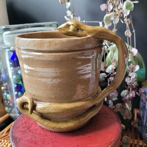 May include: A tan ceramic mug featuring a sculpted snake handle that wraps around the exterior of the vessel. The snake head forms the top of the handle, while its tail curls around the base of the mug. The surface has a smooth, glossy finish.