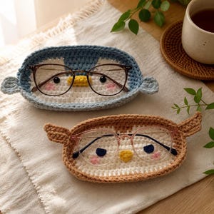 May include: Two oval-shaped crochet trays designed to resemble animal faces. The top tray is blue and white with dark-rimmed glasses, while the bottom tray is brown and white with pink-rimmed glasses. Both feature small ears and embroidered details.