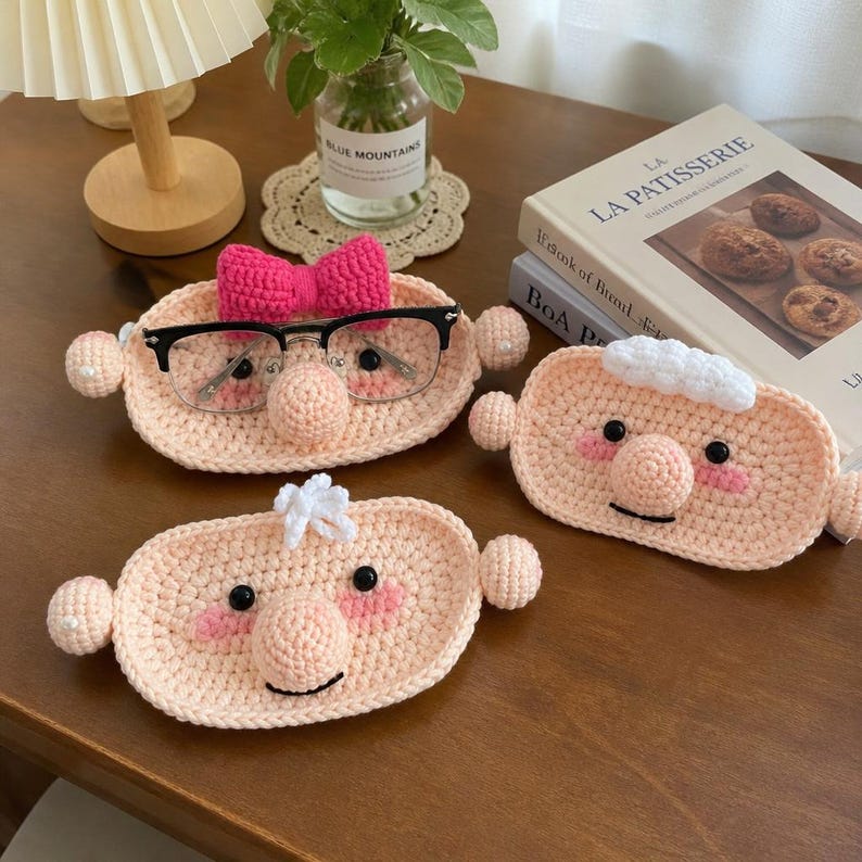 May include: Three oval-shaped crochet face trays in light peach yarn, each featuring a round nose, black bead eyes, and pink cheeks. One tray has a bright pink bow, one has white hair, and one has a small white tuft of hair on top.