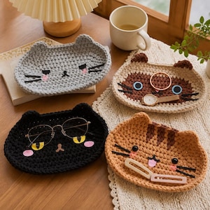 May include: Four oval-shaped crochet trays shaped like cat faces in gray, beige, black, and tan. The trays feature embroidered eyes, whiskers, and pink cheeks. The black tray holds glasses, the beige tray holds a ring and watch, and the tan tray holds hair clips.