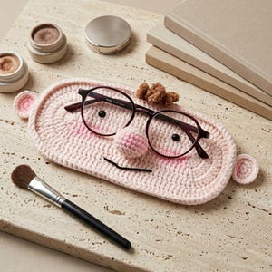 May include: A light pink oval crochet eyeglass holder featuring a small stuffed nose, two black bead eyes, and two small ears on the sides. Dark brown-rimmed glasses are resting on the holder, which also has a small brown tuft of yarn on top.