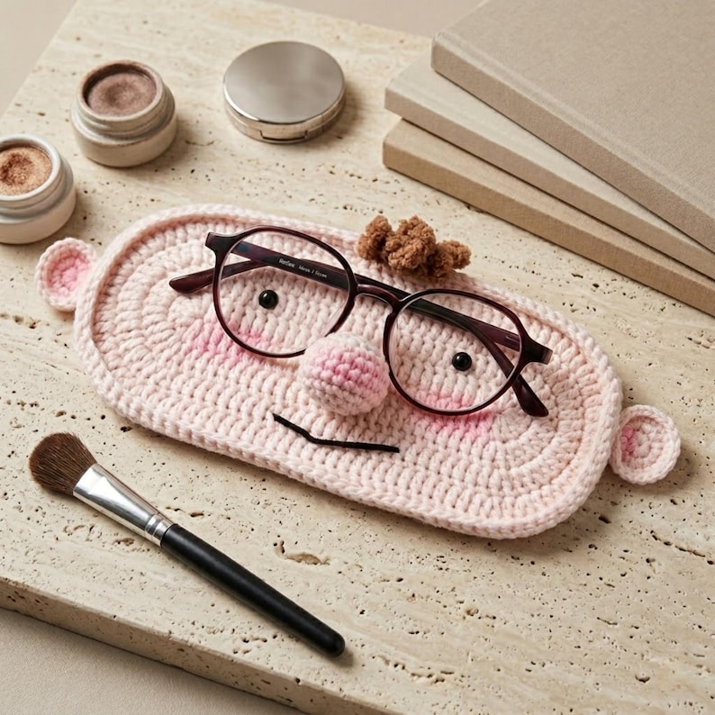 May include: A light pink oval crochet eyeglass holder featuring a small stuffed nose, two black bead eyes, and two small ears on the sides. Dark brown-rimmed glasses are resting on the holder, which also has a small brown tuft of yarn on top.