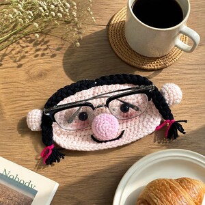 May include: A crochet eyeglass holder shaped like a face with pale pink yarn, black braided hair, and a round pink nose. The holder features a pair of black-rimmed glasses resting across the face, with small ears on the sides.