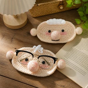 May include: Two oval-shaped, beige crochet face holders with small ears, pink cheeks, and a round nose. One holder features a tuft of white yarn on top and supports a pair of black-rimmed glasses, while the other is plain with white hair.