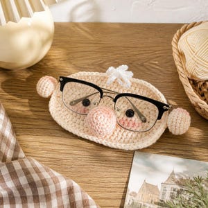 May include: A beige crochet glasses holder featuring a central round nose, two small black bead eyes, and two side spheres. The holder is shaped like a face with a small tuft of white yarn on top, designed to hold a pair of black-rimmed glasses.