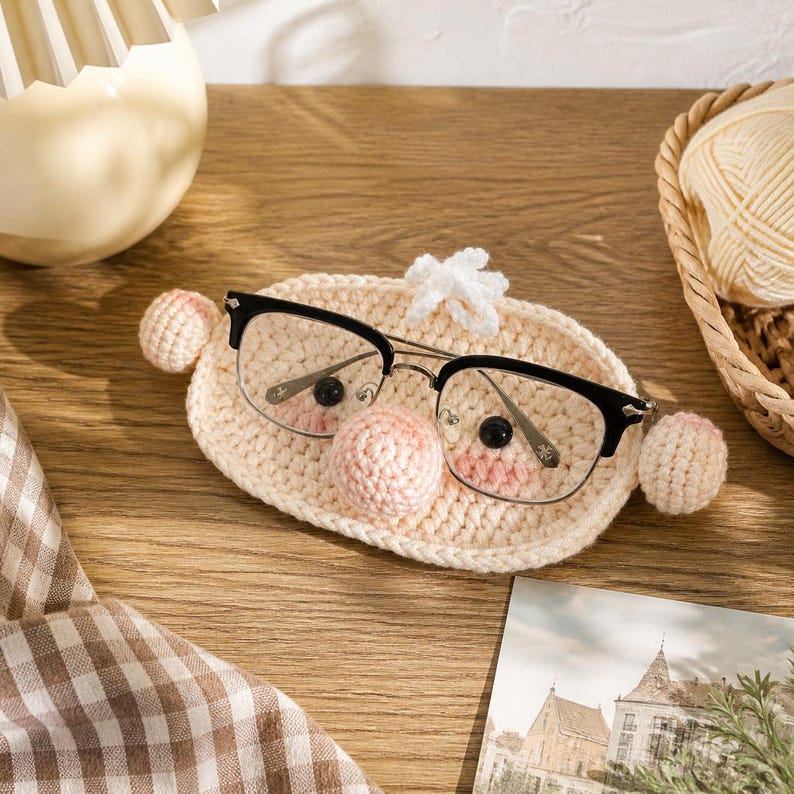 May include: A beige crochet glasses holder featuring a central round nose, two small black bead eyes, and two side spheres. The holder is shaped like a face with a small tuft of white yarn on top, designed to hold a pair of black-rimmed glasses.