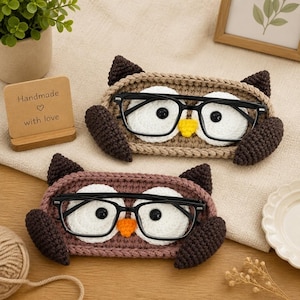 May include: Two crochet owl eyeglass holders featuring black frames resting on the owl faces. One holder is tan with a yellow beak, and the other is mauve with an orange beak. Both have dark brown wings and white circular eyes with black centers.