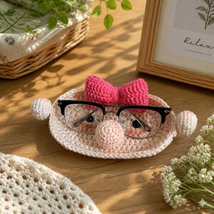 May include: A light pink crocheted oval tray featuring a bright pink bow, a central round nose-like protrusion, and two side spheres with white beads. Black-rimmed glasses rest on the tray, which is crafted from yarn.