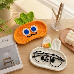May include: Two crocheted trays shaped like a carrot and a white rabbit. The orange carrot tray has green leaves and blue eyes. The white rabbit tray features long ears, a small carrot detail, and black eyes, holding black-rimmed glasses.