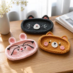 May include: Three oval crochet trays shaped like animal faces. One is dark gray with cat ears, one is light pink with rabbit ears, and one is golden brown with bear ears. A pair of black-rimmed glasses rests inside the pink tray.
