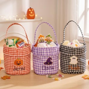 Może przedstawiać: Trzy materiałowe koszyki na Halloween w pomarańczową, fioletową i czarną kratkę gingham. Każdy koszyk ma falbankę u góry, uchwyt oraz wyhaftowany wzór: dynię dla Jamesa, kapelusz czarownicy dla Olivii i duszka dla Noaha.
