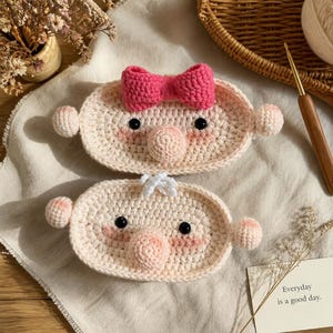 May include: Two oval-shaped crochet face scrubbies in light beige yarn. Each features a round nose, black bead eyes, and pink stitched cheeks. The top scrubby has a bright pink crochet bow, while the bottom one has a small white tuft.