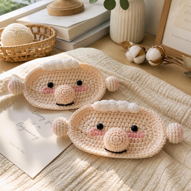 May include: Two oval-shaped, beige crochet coasters featuring a cartoon face design with white hair, black eyes, a round nose, and pink cheeks. Each coaster has small round ears on the sides and a black stitched mouth, resting on a textured fabric surface.
