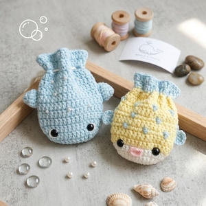 May include: Two crocheted drawstring pouches shaped like fish, one light blue with white beads and one yellow with light blue spots. Both feature black plastic eyes, side fins, and a cinched top. A white card with a whale illustration is included.