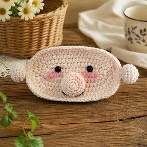 May include: A light pink oval crochet face with a prominent round nose, two small black bead eyes, and stitched pink cheeks. The piece features two small round crochet ears on the sides and a simple black stitched mouth, crafted from yarn.