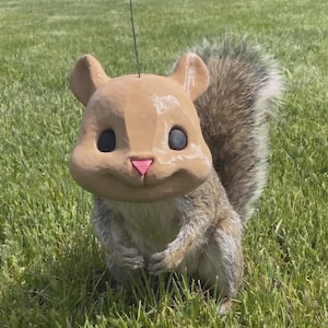 May include: A squirrel figurine featuring a large, smooth, light brown head with a small pink nose and black eyes, attached to a realistic, textured, grey and brown fur body with a bushy tail, standing upright with a thin wire extending upward.